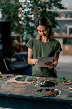 Close-up in a large green workshop, a woman with green hair near the table looks at a sketch for decoration in a tablet clipart