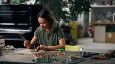 Büyük yeşil bir atölyede yeşil saçlı bir kadın onları lehim demiriyle el yapımı yuvarlak bir aynaya seren dekoratif çiçekler yaratıyor..