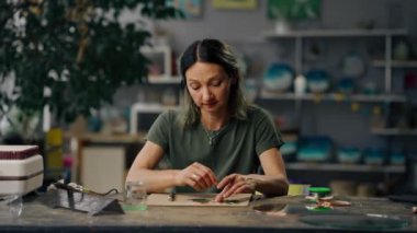 Büyük yeşil bir atölyede yeşil saçlı bir kadın onları lehim demiriyle el yapımı yuvarlak bir aynaya seren dekoratif çiçekler yaratıyor..