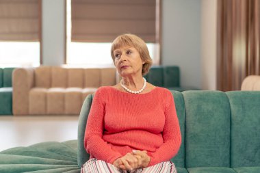 Modern bir klinikte, bekleme odasında, mercan kazaklı yaşlı bir kadın yumuşak yeşil kadife bir koltukta oturuyor ve doktorun aramasını bekliyor.