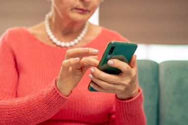 Bekleme odasındaki modern bir klinikte, mercan kazaklı yaşlı bir kadın yeşil kadife koltukta oturuyor, doktorun aramasını bekliyor, telefonundan haberleri okuyor.