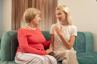 Modern bir klinikte, bekleme odasında, mercan kazaklı yaşlı bir kadın yeşil bir kanepede oturuyor ve torunu el ele tutuşup, doktorun aramasını bekliyor.