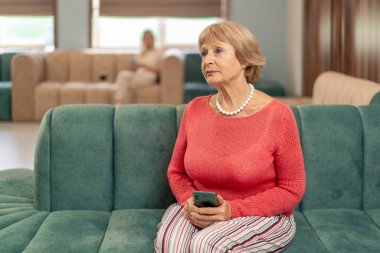 Modern bir klinikte, bekleme odasında, mercan kazaklı yaşlı bir kadın yeşil kadife koltukta oturuyor, doktorun telefonla aramasını bekliyor.