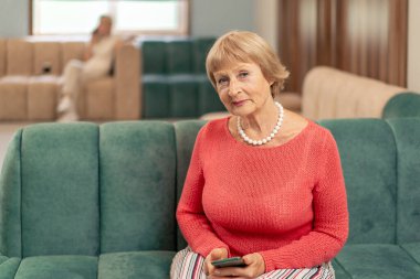 Modern bir klinikte, bekleme odasında, mercan kazaklı yaşlı bir kadın yeşil kadife koltukta oturuyor, doktorun telefonla aramasını bekliyor.