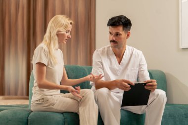 Modern bir klinikte, bekleme odasında, açık elbiseli genç bir kız kanepede bir doktorla oturuyor ve sağlık sorununu konuşuyor.