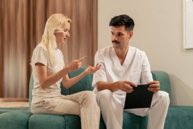Modern bir klinikte, bekleme odasında, açık elbiseli genç bir kız kanepede bir doktorla oturuyor ve sağlık sorununu konuşuyor.