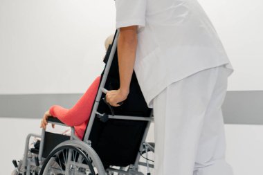 Modern bir klinikte, beyaz bir koridorda, tekerlekli sandalyede mercan süveter giyen yaşlı bir kadın sınırlı bir hareketle, hemşirelerin hareket kabiliyetini kısıtlıyor.