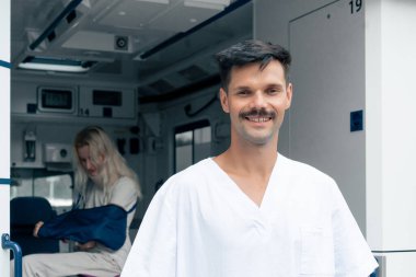 Sokakta bir ambulansta, boynu ve kolu yaralı genç bir hasta bir hastayı kliniğe naklediyor, genç bir doktor da arabanın önünde.