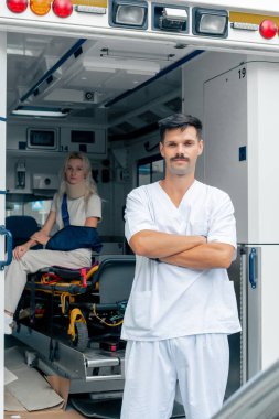 Sokakta bir ambulansta, boynu ve kolu yaralı genç bir hasta bir hastayı kliniğe naklediyor, genç bir doktor da arabanın önünde.