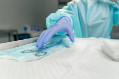 Modern klinikte, parlak, geniş bir ameliyathanede steril eldivenli bir doktor paketleri açmak için cerrahi klemp alır ve...