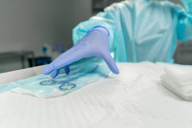 Modern klinikte, parlak, geniş bir ameliyathanede steril eldivenli bir doktor paketleri açmak için cerrahi klemp alır ve...