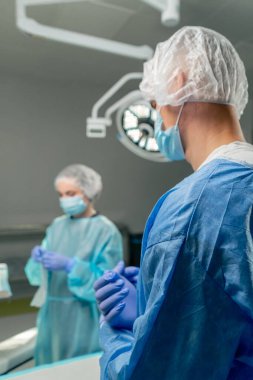 Modern bir klinikte, parlak, geniş bir ameliyathanede, bir doktor, steril eldivenli bir cerrah, paketsiz bir cerrahi kıskacı alır ve takım çalışması için asistanına verir.