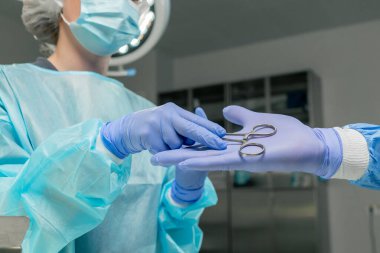 Modern bir klinikte, parlak, geniş bir ameliyathanede, steril eldivenli bir doktor, boş bir cerrahi kelepçeyi alır ve takım çalışması için asistanına verir.