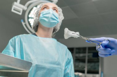 Modern bir klinikte, parlak, geniş bir ameliyathanede steril eldivenli bir doktor takım çalışması için temizleme çubuğu için cerrahi klemp tutuyor.