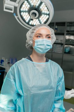 Modern bir klinikte, parlak, geniş bir ameliyathanede steril eldivenli bir doktor temizlik, takım çalışması için bir pens tutuyor, bir hemşire cerrahı izliyor.