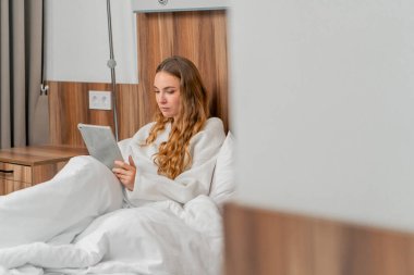 Modern bir klinikte, parlak bir VIP odasında, ameliyattan önce beyaz önlüklü genç bir kız yatağa uzanıyor, tableti karıştırıyor, seçeneklere bakıyor.