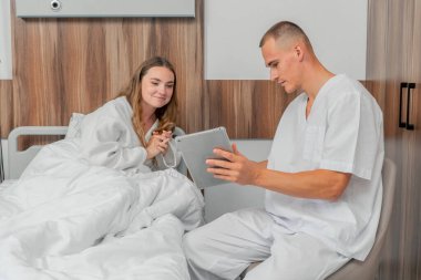 Modern bir klinikte, parlak bir VIP odasında beyaz önlüklü genç bir kız hasta seçiminden önce yatağında bir doktora tabletle danışarak yatıyor.