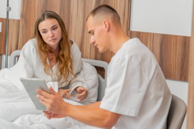 Modern bir klinikte, parlak bir VIP odasında beyaz önlüklü genç bir kız hasta seçiminden önce yatağında bir doktora tabletle danışarak yatıyor.