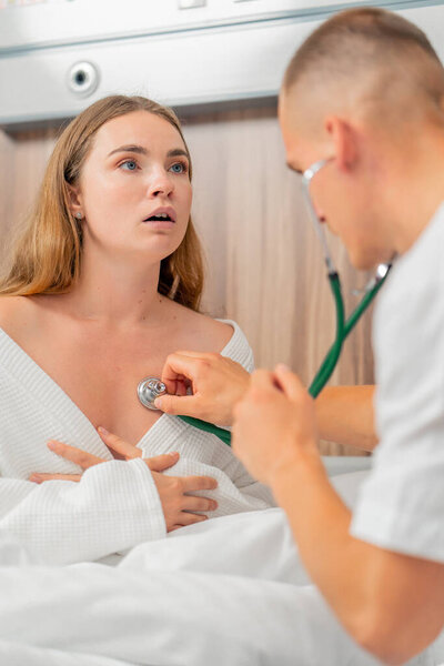 in a modern clinic in a bright VIP room, a young girl in a white coat lies on the bed sick consultation with a doctor, the doctor listens to the lungs with a stethoscope