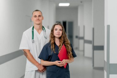 Kliniğin beyaz koridorunda mavi üniformalı bir kadın doktor ve beyaz üniformalı bir doktor kameraya bakar, takım arkadaşı çalışır.