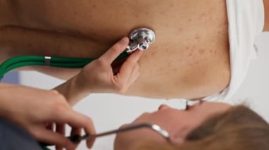 Modern klinikte, küçük terapistin ofisindeki dikey videoda, steteskoplu genç bir kız doktor, genç bir hastayla randevu ayarlıyor, bir sorunu teşhis ediyor, akciğerleri dinliyor.
