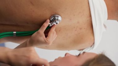 Modern klinikte, küçük terapistin ofisindeki dikey videoda, steteskoplu genç bir kız doktor, genç bir hastayla randevu ayarlıyor, bir sorunu teşhis ediyor, akciğerleri dinliyor.