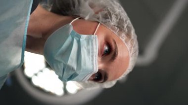 Modern bir klinikte, parlak, geniş bir ameliyathanede, dikey bir video, steril eldivenli bir doktor temizlik, takım çalışması için bir bez çubuğu tutuyor, bir hemşire de...