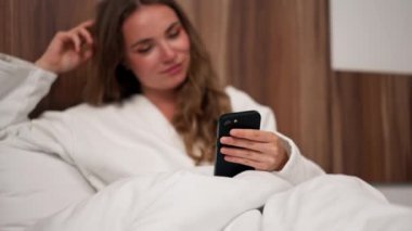 Modern bir klinikte parlak bir VIP odasında beyaz önlüklü genç bir kız telefonda estetik ameliyat için bekliyor mutlu hasta implantları seçiyor.