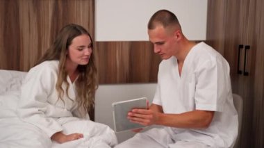 Modern bir klinikte, parlak bir VIP odasında beyaz önlüklü genç bir kız hasta seçiminden önce yatağında bir doktora tabletle danışarak yatıyor.
