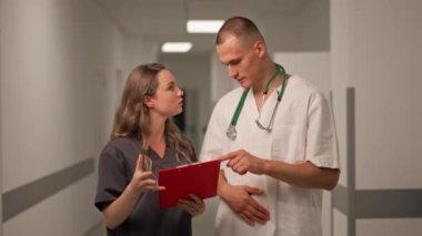 Kliniğin beyaz koridorunda mavi üniformalı kadın doktor ve beyaz üniformalı bir doktor hastanın sonuçlarını kırmızı klasörde tartışıyor.