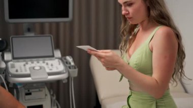 Kliniğe yakın çekim, jinekoloji ofisine yakın çekim, genç bir hasta koltukta oturuyor, genç bir kız, bir jinekolog, bir erkek, ona hamilelik hakkında bilgi veriyor, ultrason görüntüsü gösteriyor.