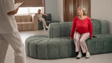 Modern bir klinikte, bekleme odasında, mercan kazaklı yaşlı bir kadın ayakta duruyor, bir doktor sonuçlarla yaklaşıyor, iletişim kuruyor, iyi sonuçlar veriyor.