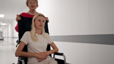 Kliniğe yakın beyaz bir koridorda mercan kazaklı yaşlı bir kadın hareket kabiliyeti sınırlı olan tekerlekli sandalyede torununu taşıyor.