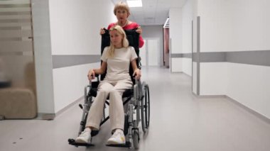 Klinikte beyaz bir koridorda mercan kazaklı yaşlı bir kadın hareket kabiliyeti sınırlı olan tekerlekli sandalyede torununu taşıyor.