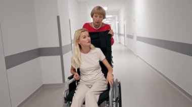 Klinikte beyaz bir koridorda mercan kazaklı yaşlı bir kadın hareket kabiliyeti sınırlı olan tekerlekli sandalyede torununu taşıyor.