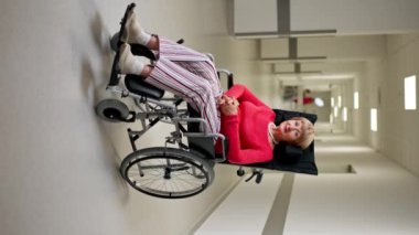 Modern bir klinikte dikey video beyaz bir koridorda mercan kazaklı yaşlı bir kadın bebek arabasında sınırlı bir hareketle pozitif bir tutum sergiliyor.