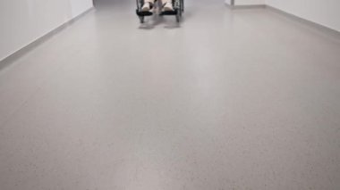 Kliniğe yakın beyaz bir koridorda mercan kazaklı yaşlı bir kadın hareket kabiliyeti sınırlı olan tekerlekli sandalyede torununu taşıyor.