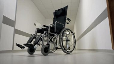 Modern bir klinikte, beyaz bir koridorda engelliler için yeni bir siyah sandalye, hareket yardımı, modern tedavi yöntemleri var.