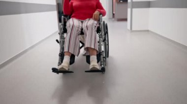 Modern bir klinikte, beyaz bir koridorda, tekerlekli sandalyede mercan süveter giyen yaşlı bir kadın sınırlı bir hareketle, hemşirelerin hareket kabiliyetini kısıtlıyor.