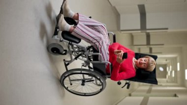Modern bir klinikte beyaz bir koridorda dikey bir video, bebek arabasıyla mercan kazak giyen yaşlı bir kadın telefonda haberleri okuyan olumlu bir kişi.
