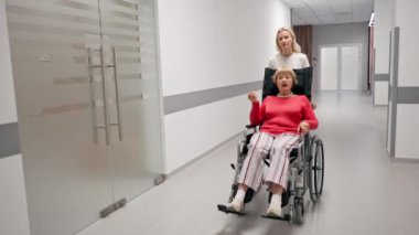 Modern bir klinikte, beyaz bir koridorda, tekerlekli sandalyede mercan süveter giyen yaşlı bir kadın sınırlı bir hareketle, hemşirelerin hareket kabiliyetini kısıtlıyor.