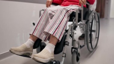 Modern bir klinikte, beyaz bir koridorda, tekerlekli sandalyeli, mercan kazaklı yaşlı bir kadın, sınırlı hareketli, hasta bakıcı hareketlerine yardımcı oluyor.