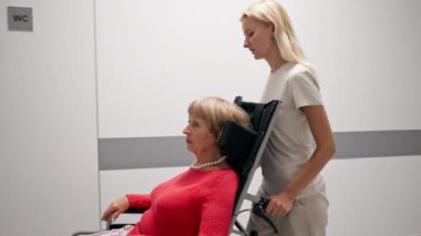 Modern bir klinikte, beyaz bir koridorda, tekerlekli sandalyeli, mercan kazaklı yaşlı bir kadın, sınırlı hareketli, hasta bakıcı hareketlerine yardımcı oluyor.