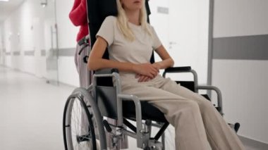 Klinikte beyaz bir koridorda mercan kazaklı yaşlı bir kadın hareket kabiliyeti sınırlı olan tekerlekli sandalyede torununu taşıyor.