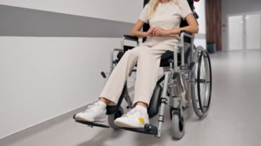 Kliniğe yakın beyaz bir koridorda mercan kazaklı yaşlı bir kadın hareket kabiliyeti sınırlı olan tekerlekli sandalyede torununu taşıyor.