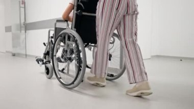 Kliniğe yakın beyaz bir koridorda mercan kazaklı yaşlı bir kadın hareket kabiliyeti sınırlı olan tekerlekli sandalyede torununu taşıyor.