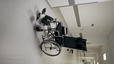 Beyaz bir koridordaki modern bir klinikte, engelliler için yeni bir siyah sandalye, hareket yardımı ve modern tedavi yöntemleri bulunmaktadır.