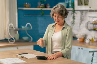 Parlak bir mutfakta açık yeşil tişörtlü yaşlı bir kadın, içinde para olan bir masanın yanında duruyor. Birikimleri sayıyor.