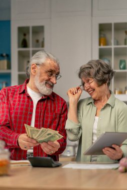 Parlak bir mutfakta yaşlı, sevimli bir çift masalarında aylık ödeme faturalarını sayıyor gülümseyerek tablete bakıp gülüyor.