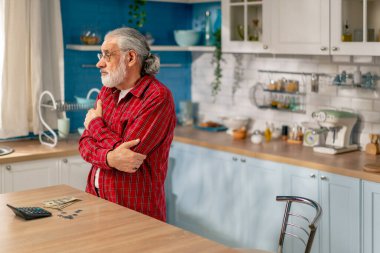 Aydınlık bir mutfakta masanın üstünde kırmızı gömlekli yaşlı bir adam masraflarını aylık fatura olarak sayar.